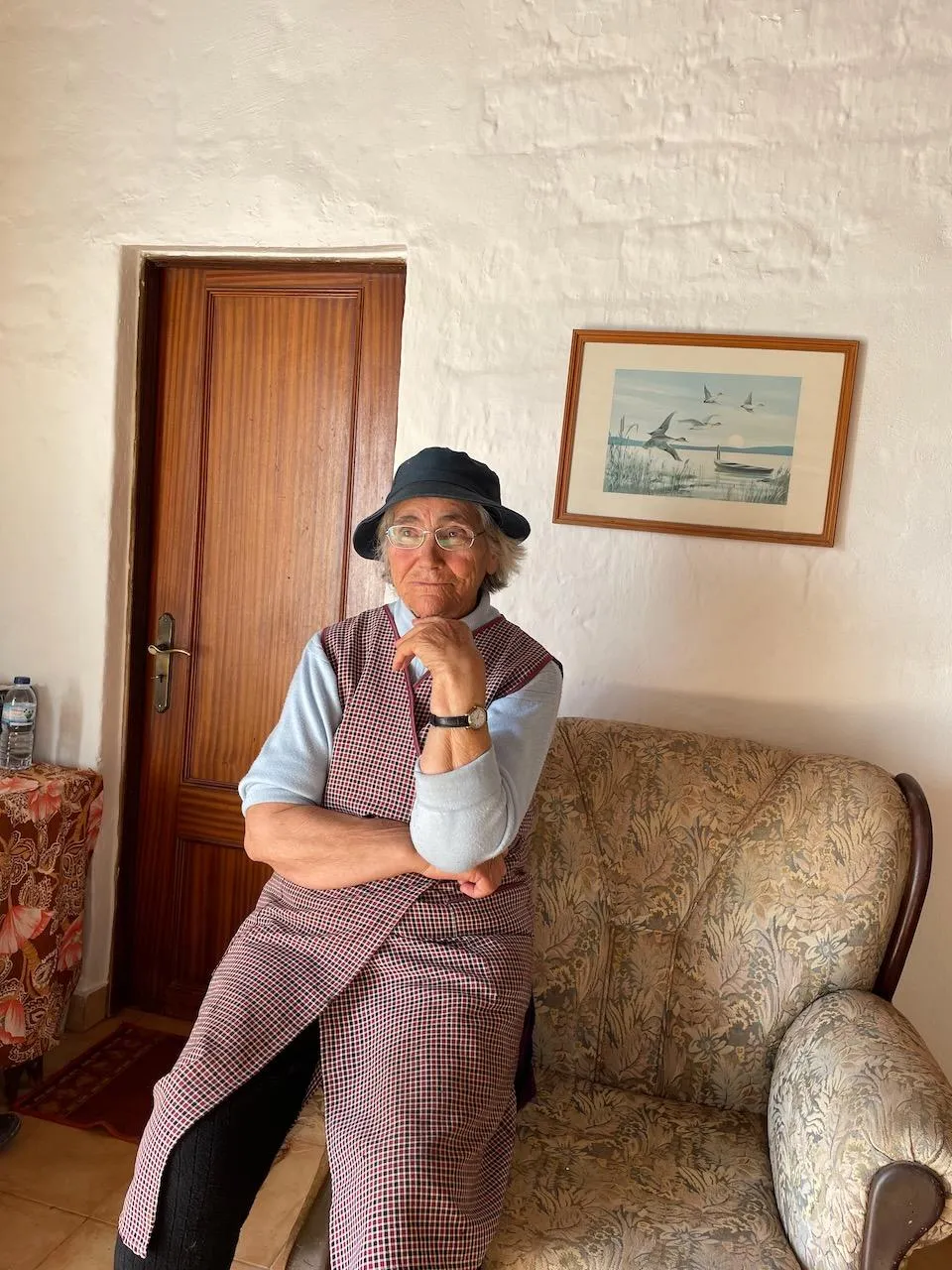 Sofia, a woman in a black hat and checked apron, sitting on a floral sofa in her living room